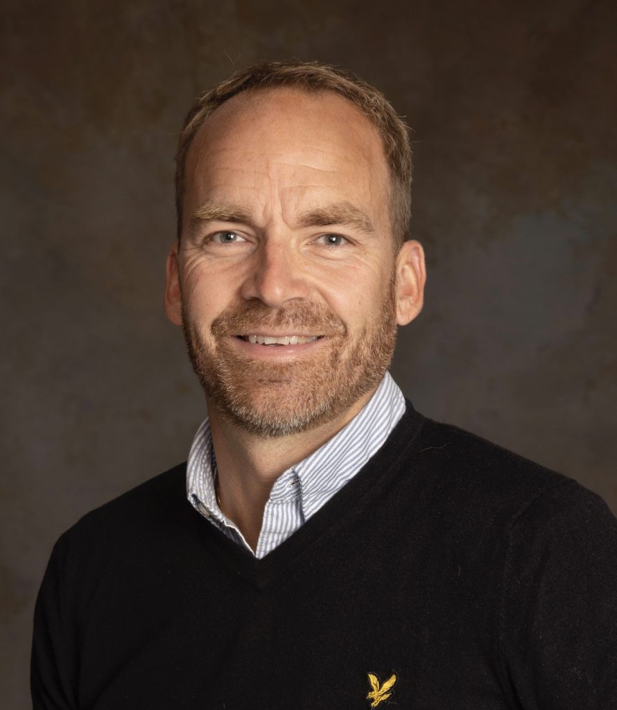 A man with short light brown hair and a beard, wearing a black sweater over a white collared shirt, smiles in front of a brown mottled background.